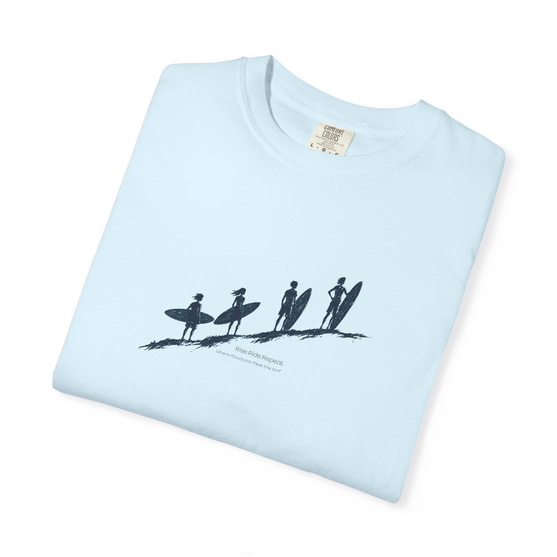A light blue folded t-shirt with a black graphic of four surfers on a beach and the text 'Suna Tide Surfing' printed on the front, set against a plain white background.