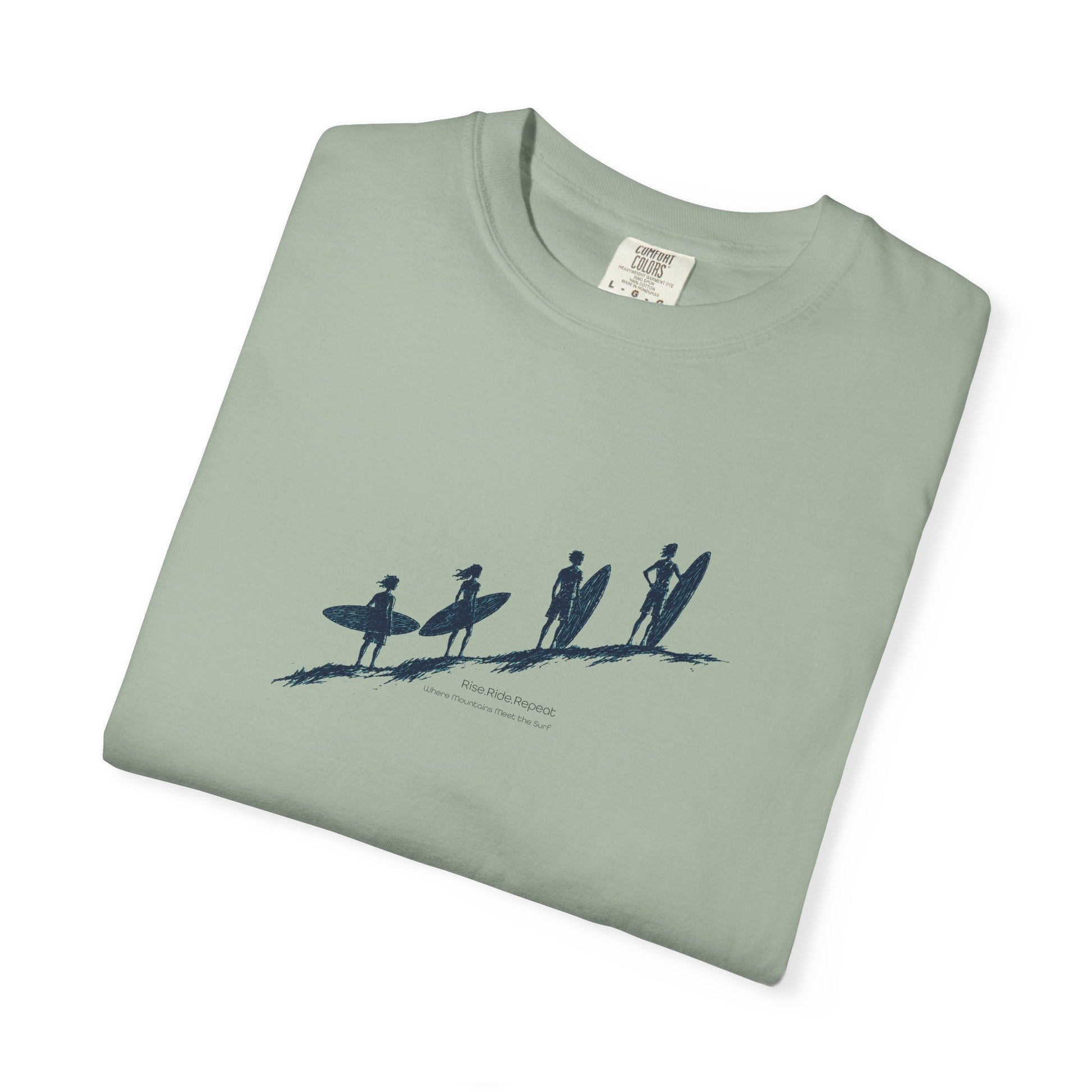 A folded sage green t-shirt with a dark blue graphic of four surfers on a beach, displayed on a clean white background.