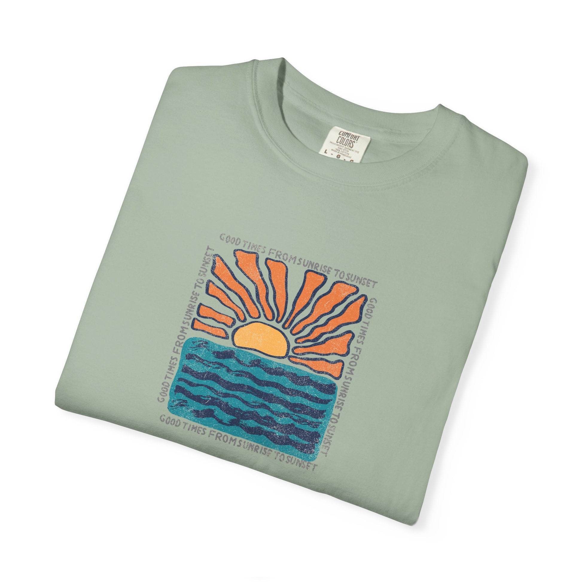 A folded sage green t-shirt with a colorful graphic of a rising sun over blue waves, with the text 'GOOD TIMES FROM THE SUNNY SIDE OF THE MOUNTAIN' printed around the design.